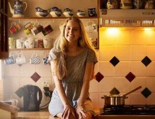 A girl sitting in kitchen near clean stove is happy with Kitchen Cabinet Cleaning service is smiling with 10's of tea mugs behind