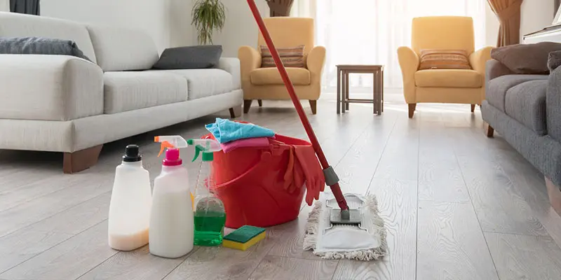 Cleaning tools including spray bottles, gloves, mop, and bucket placed on a wooden floor in a clean, modern living room