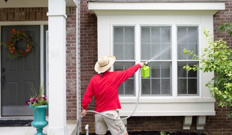 Exterior Window Washing for Southampton Homes A person using a hose and cleaner to wash exterior windows on a brick house in Southampton, addressing coastal salt and grime buildup.