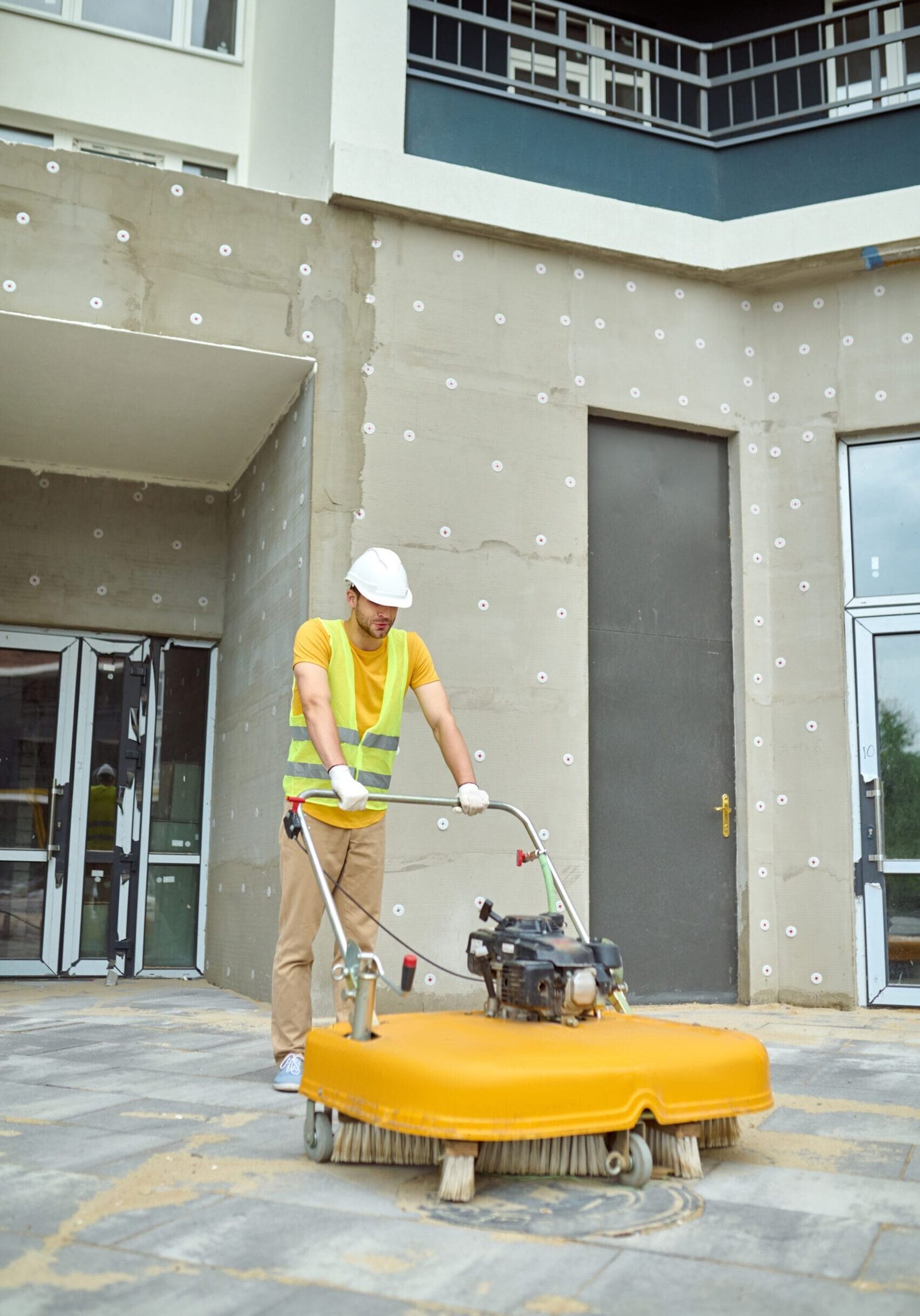 Cleaning. Man in protective helmet and gloves operating special equipment with brush cleaning construction site in new housing complex outdoors