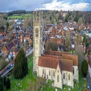 Aerial view of the historic town of Farnham, England, featuring the iconic spire of St. Andrew's Church rising above the quaint mix of red-tile roofed buildings, cobblestone streets, and surrounding greenery.