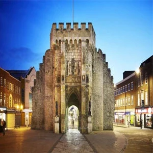 A historic stone gatehouse in Southampton, UK, illuminated at night with shops and pedestrians visible in the background.